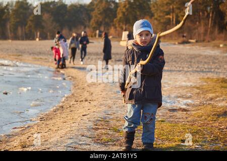 Small four years caucasian boy standing near riverside and holding long wooden stick looking at camera in sunny winter day before sunset Stock Photo