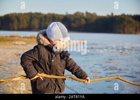 Small four years caucasian boy standing near riverside and playing with long wooden stick in sunny winter day Stock Photo