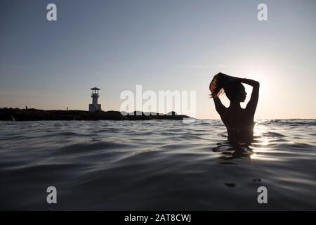 Rear view of woman bathing in the ocean at sunset, lighthouse in the distance. Stock Photo
