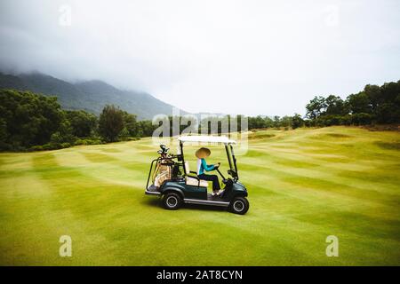 Woman wearing traditional straw hat driving golf cart on the green of a golf course under a cloudy sky. Stock Photo