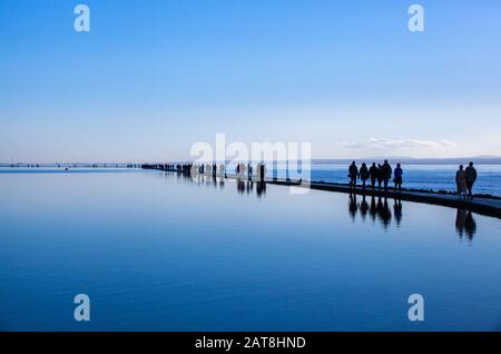 People walking around the Marine Lake at West Kirby on the Wirral Peninsula in winter on the wall path between the boating lake and the Dee estuary Stock Photo
