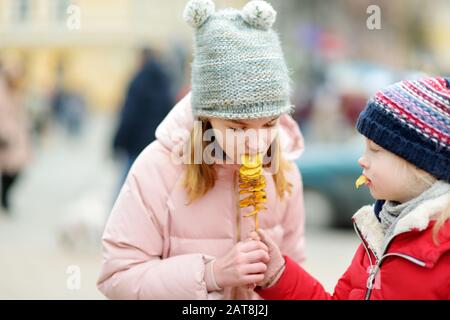 Cute little girls eating fried potato on a stick on cold winter day outdoors. Snacks for kids. Stock Photo