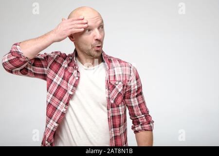 Funny, shocked bald man, with a bizarre expression on face, holds hand near head and looks surprised to the side. Copy space. Stock Photo