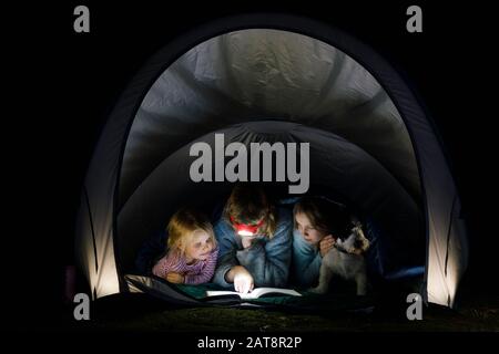 Girls laying in a camping tent wrapped in a sleeping bag with dog reading a book with flashlight at night during summer holidays Stock Photo