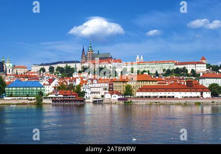Prague Castle and St. Vitus Cathedral with many small houses under red roofs near Vltava river against vibrant blue sky and white clouds in daytime. P Stock Photo