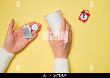 Mans hands with tea bag, white paper cup and red alarm clock on yellow background. Top view. Concept of break, tea time. Stock Photo