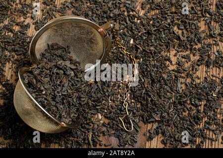 Black tea in open strainer with chain on scattered tea on wooden background. Concept of a tea shop. Stock Photo