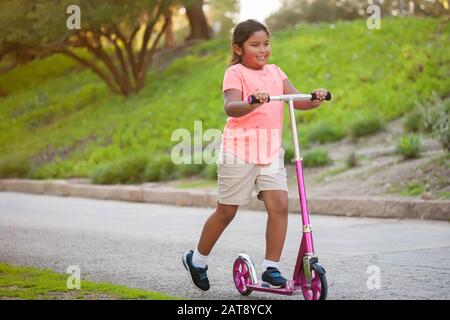 A little girl riding a pink scooter with confidence in a street lined with trees and grass. Stock Photo