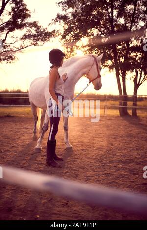 Woman in equestrian gear petting a horse while looking away at the sunset. fun on countryside,  golden hour. Freedom nature concept. Stock Photo