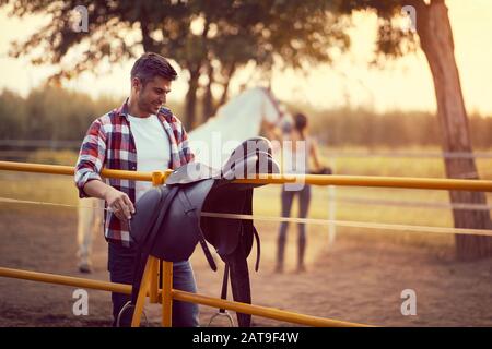 Man preparing a horse saddle for a ride,woman with a horse in the back. Training  on countryside, sunset golden hour. Freedom nature concept. Stock Photo