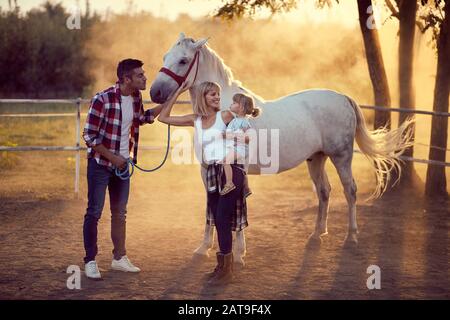 Happy parents with their daughter on the horse ranch, family concept .Young happy family having fun at countryside outdoors. Sunset, golden hour