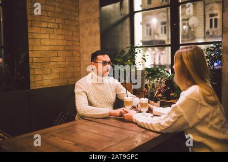 Young happy couple in love in cafe, man and woman together smile hugging, drinking coffee. Couple Talking In Cafe In Winter. Happy couple enjoying a Stock Photo