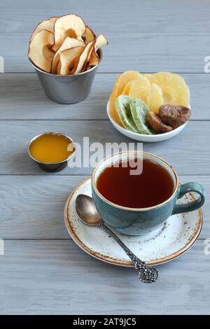 Vegetarian dessert: pieces of candied pineapple, kiwi and mango, dried figs, honey, apple chips in small bowls and a cup of black tea on a gray wooden Stock Photo
