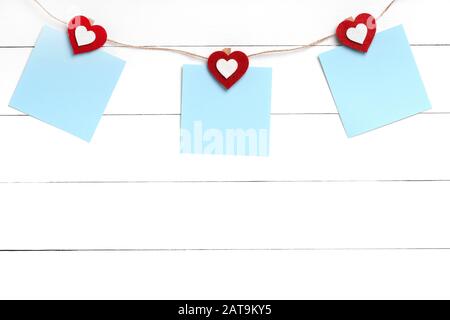 Set of three blank stickers hanging on string with clothespins behind white wooden surface. Paper notes held on rope with red hearts. Copy space, top Stock Photo