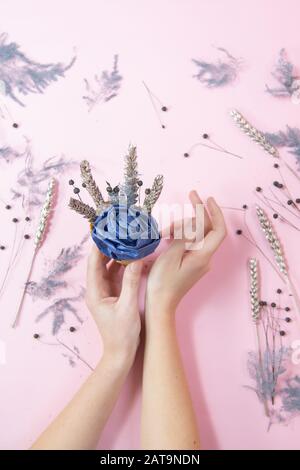 holiday celebration woman hands holding waffle cone blue handmade rose with dried flowers,copy space pink color paper minimalist background flat lay. Stock Photo