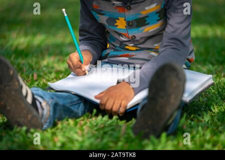 Little child schoolkid writing on a notebook with pencil while sitting on green grass in the park. Kid education concept. Stock Photo