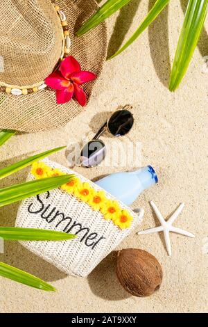 Small bag with word SUMMER, straw hat, coconut, sunglasses and palm leafs on sand, top view, vertical composition Stock Photo