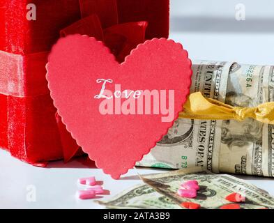 A red heart and a read gift box with another box wrapped in money and bills and candy in the foreground. the cost of love Stock Photo
