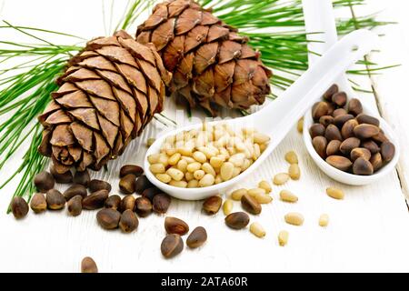 Pine nuts in two spoons, two cedar cones and green branches on a wooden board background Stock Photo