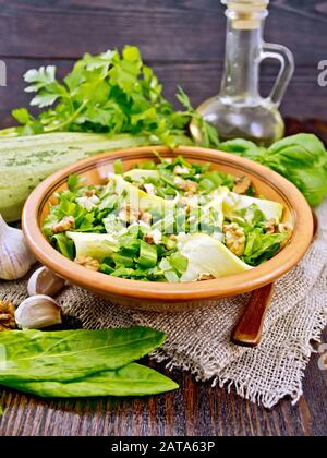 Salad of young zucchini, sorrel, garlic and nuts, seasoned with vegetable oil in clay plate on napkin of sackcloth on a dark wooden board background Stock Photo