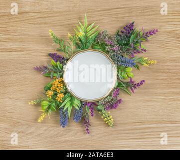 Summer composition of meadow flowers and an empty white porcelain plate with a gold rim on a wooden tabletop background Stock Photo