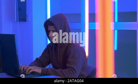 African hacker dressed in hoodie working on the computer in his secret place with warm neon lights Stock Photo