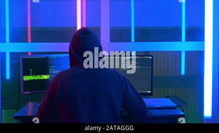 Back view of hacker in a black jacket and a hood on his head writing hacker programs. He working on personal computer in hacker dark room. Stock Photo