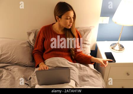 Student girl working on laptop on bed distracted by phone at the night time Stock Photo