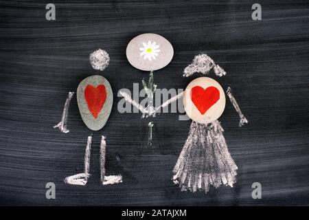 Pebbles with red hearts inside chalk drawing of man and woman holding flower in hands on blackboard. Close-up. Stock Photo