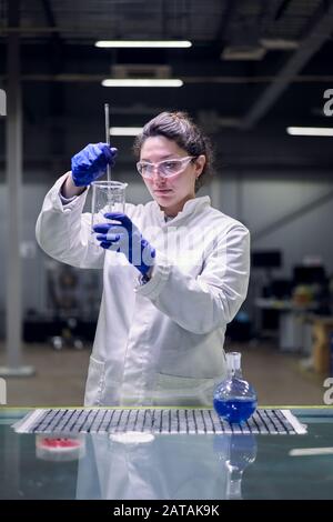 Serious lab girl in glasses and white coat with experimental glass in her hands conducts experiments on defocused background Stock Photo