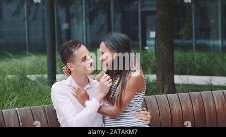 Young multiethnic adults stroking each other and talking on date outdoors. Black woman sitting on man's knees. Stock Photo