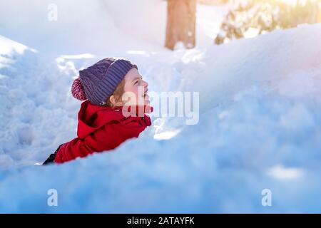 Joyful child in the snow, cute cheerful little girl lying down in the pile of snow and laughing, enjoying winter holidays, happy carefree childhood