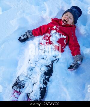 Little girl playing snow angel, cheerful child lying down in the snow and screaming of joy, outdoor fun, enjoying winter holidays, happy childhood Stock Photo