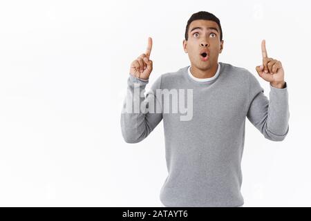 Speechless and curious, excited hispanic masculine guy, standing startled and astonished as pointing fingers up folding lips and say wow pumped over s Stock Photo