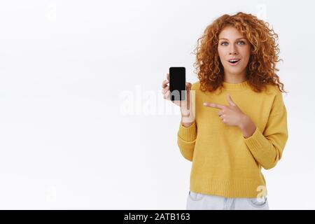 Amused, charming ginger girl with curly hair in yellow sweater, holding smartphone, pointing mobile screen and smiling amazed, impressed using cool Stock Photo