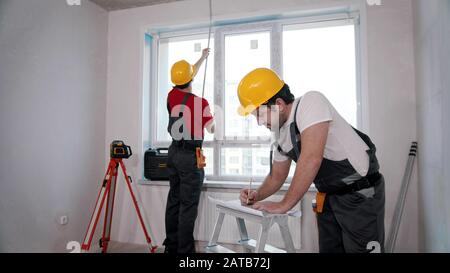 Apartment repair in the room - two men workers measuring the distance from the windowsill to the ceiling and another writing notes about it Stock Photo
