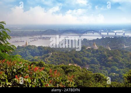 Panoramic view of Buddhist Temples on the side of the Irrawaddy River, surrounded by green vegetation, against a clear blue sky covered by white cloud Stock Photo