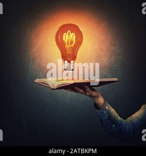 Male student hand holding an open book with a shining light bulb over a dark blackboard background. Symbol of knowledge, intelligence and brilliant id Stock Photo