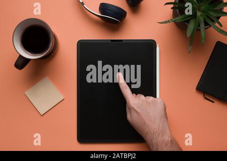 Stock photo of a young man's hand pointing the screen of a white tablet and some desk objects on a coral background Stock Photo