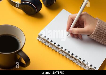 Stock photo of an open notebook page with a hand writing in it, coffee and headphones on a yellow background Stock Photo