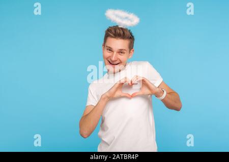 Portrait of happy angelic man with halo above head making heart shape with hands and smiling to camera, sharing romantic amorous feelings, declaration Stock Photo