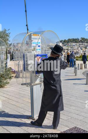 An orthodox jew man is calling from a public payphone in Jerusalem, Israel. Dressed with a hasidic garb. In front of him the view over hills of East Stock Photo