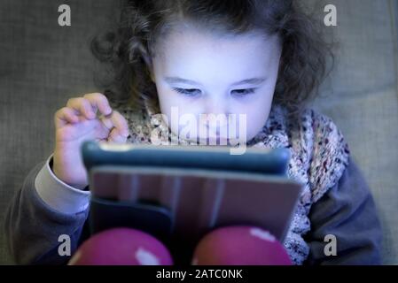 Little girl uses a digital tablet. Two-year-old child looks electronic device at home. Cute toddler sits on a couch and plays computer games. Face of Stock Photo