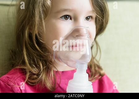 Little girl with inhaler mask close-up. Sick kid breathes through a nebulizer at home. Face of toddler using equipment to treat asthma or bronchitis. Stock Photo