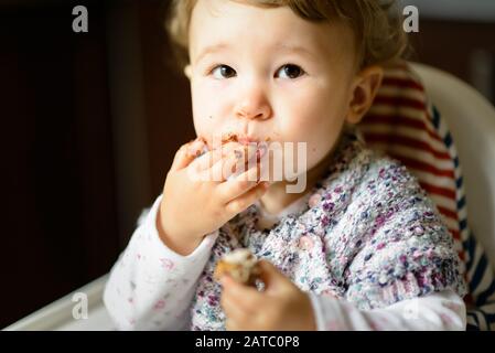 Eating baby girl with messy face. The one-year child eats and puts fingers in his mouth. Stock Photo