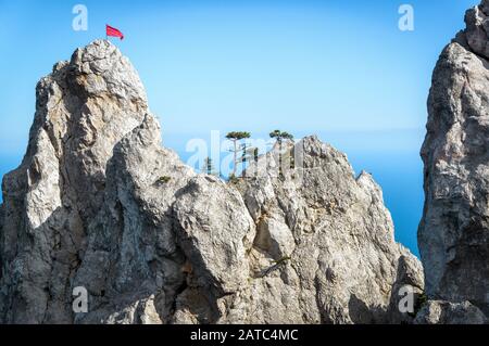 The rock on Mount Ai-Petri with a red flag and trees over Black Sea in Crimea, Russia. Ai-Petri is one of the highest mountains in Crimea and tourist Stock Photo