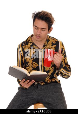 A tall young man sitting on a chair in the studio, reading his book and holding a mug with coffee, isolated for white background Stock Photo