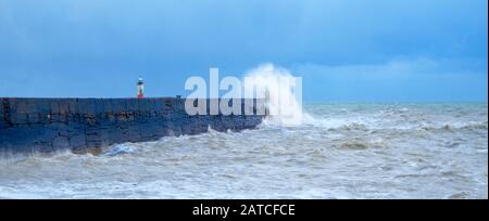 A harbor wall with a rough stormy sea crashing against the wall causing the sea to be blurred and in motion, behind is a red and white lighthouse, wav Stock Photo