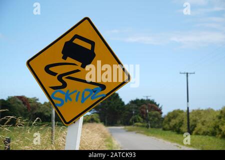 Yellow and black vehicle skid warning road sign with skidz sprayed in blue graffiti against a blue sky and green countryside  rural background rural Stock Photo