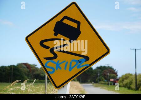 Yellow and black vehicle skid warning road sign with skidz sprayed in blue graffiti against a blue sky and green countryside  rural background rural Stock Photo